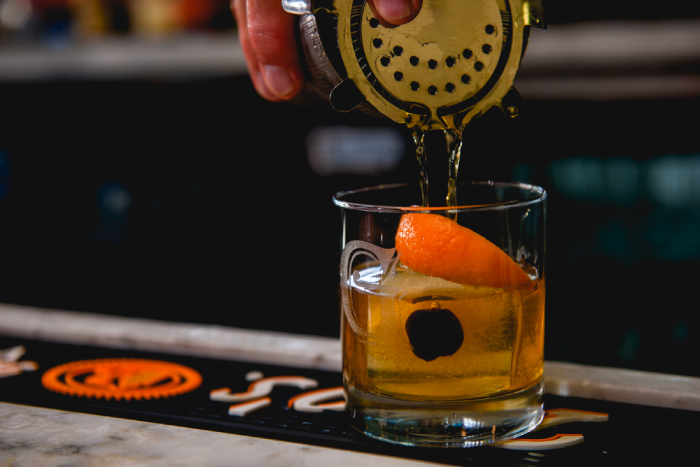 Hand pouring cocktail into a glass with orange peel.