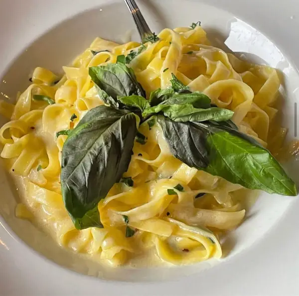 A white bowl of fresh fettuccine pasta with a creamy alfredo sauce, garnished with fresh basil leaves.
